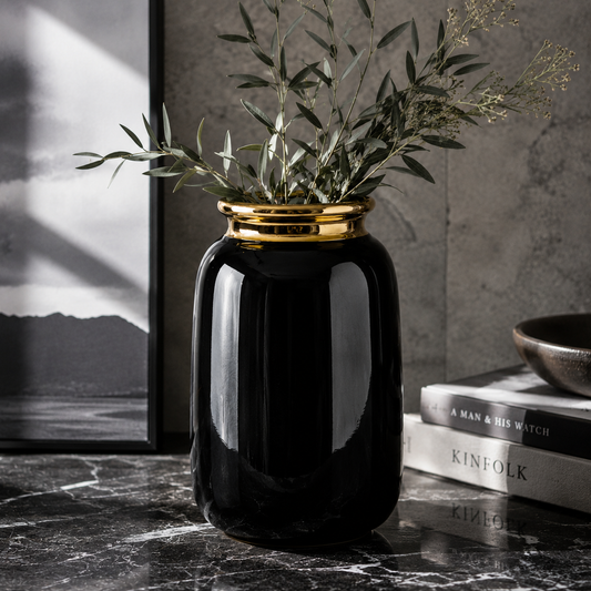 Black vase with gold rim and greenery on a dark marble surface with books in the background