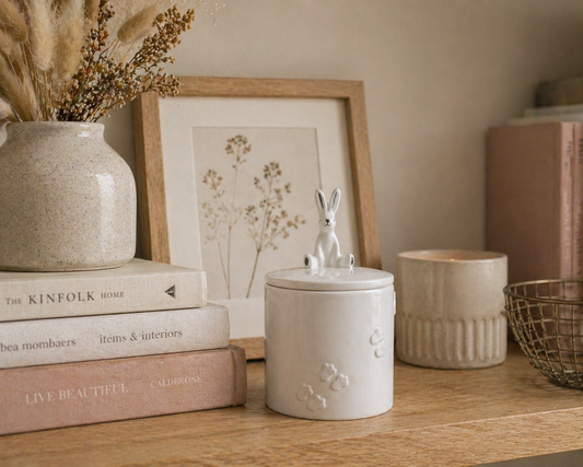 A white ceramic lidded jar with a small rabbit figure on top, styled on a wooden shelf next to a beige candle, a framed botanical print, and books.