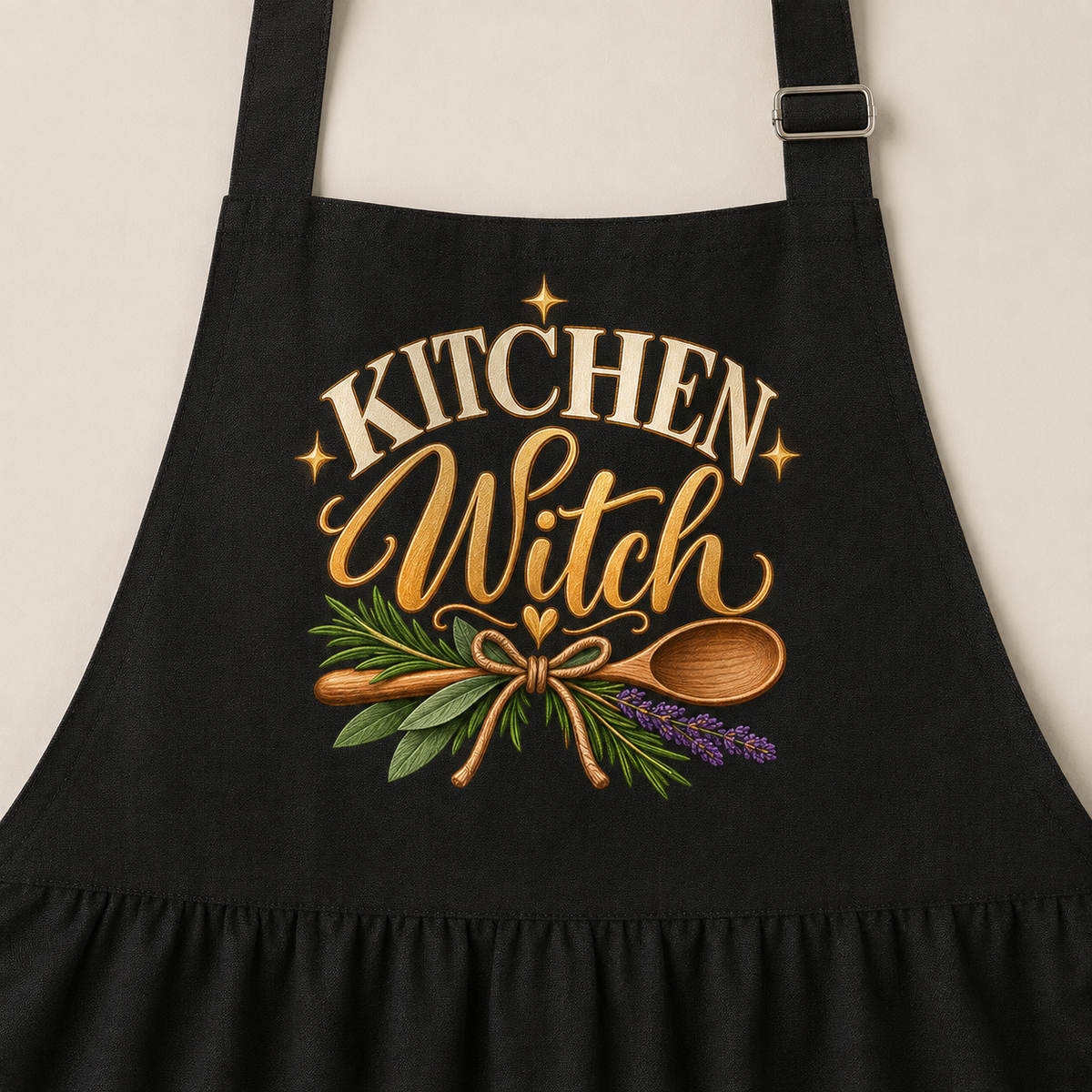 A black kitchen apron with a ruffled hem, featuring a graphic print that reads 'KITCHEN Witch' in gold lettering, decorated with a wooden spoon, green herbs, and purple lavender sprigs.