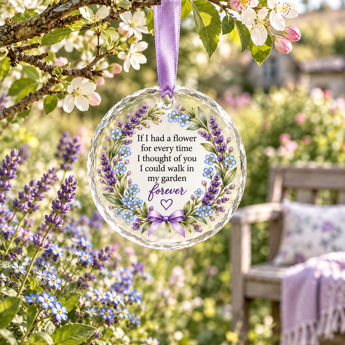 A round clear glass memorial ornament hanging from a purple ribbon, printed with a floral wreath design and the message 'If I had a flower for every time I thought of you I could walk in my garden forever.' The ornament is photographed outdoors among blooming flowers and a garden bench, with no person present.
