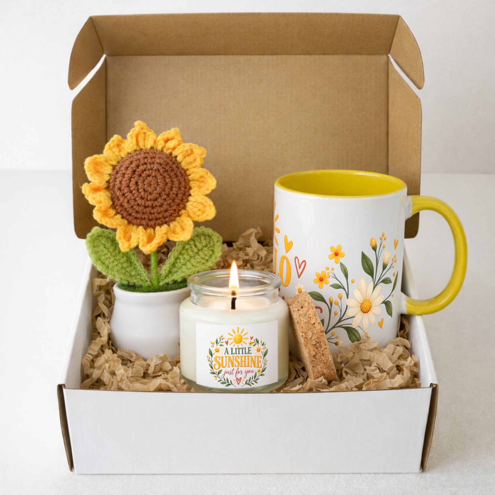 A gift box set containing a crocheted sunflower, a white candle with a 'A Lime Sunshine' label, a white and yellow ceramic mug with floral patterns, and a small cork coaster, all arranged in brown paper shred inside an open white cardboard box.