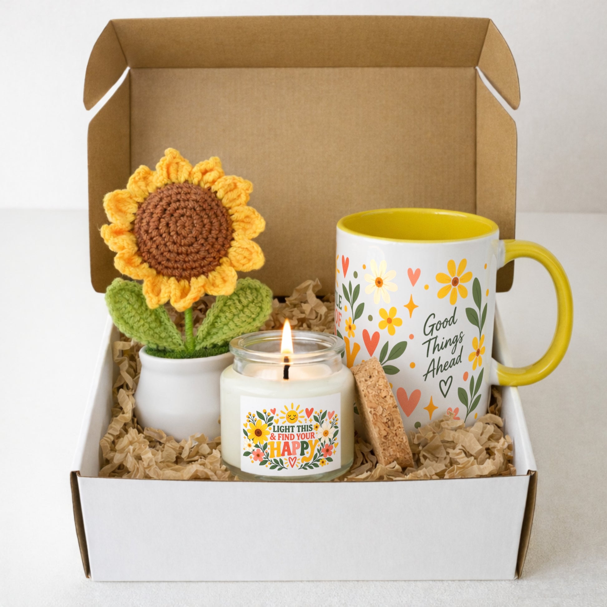 A gift box set containing a crocheted sunflower in a small white pot, a white candle in a glass jar with a yellow and multicolor label, a ceramic mug with a yellow handle and rim, and a small cork coaster, all arranged in brown paper shred inside an open white and brown gift box.