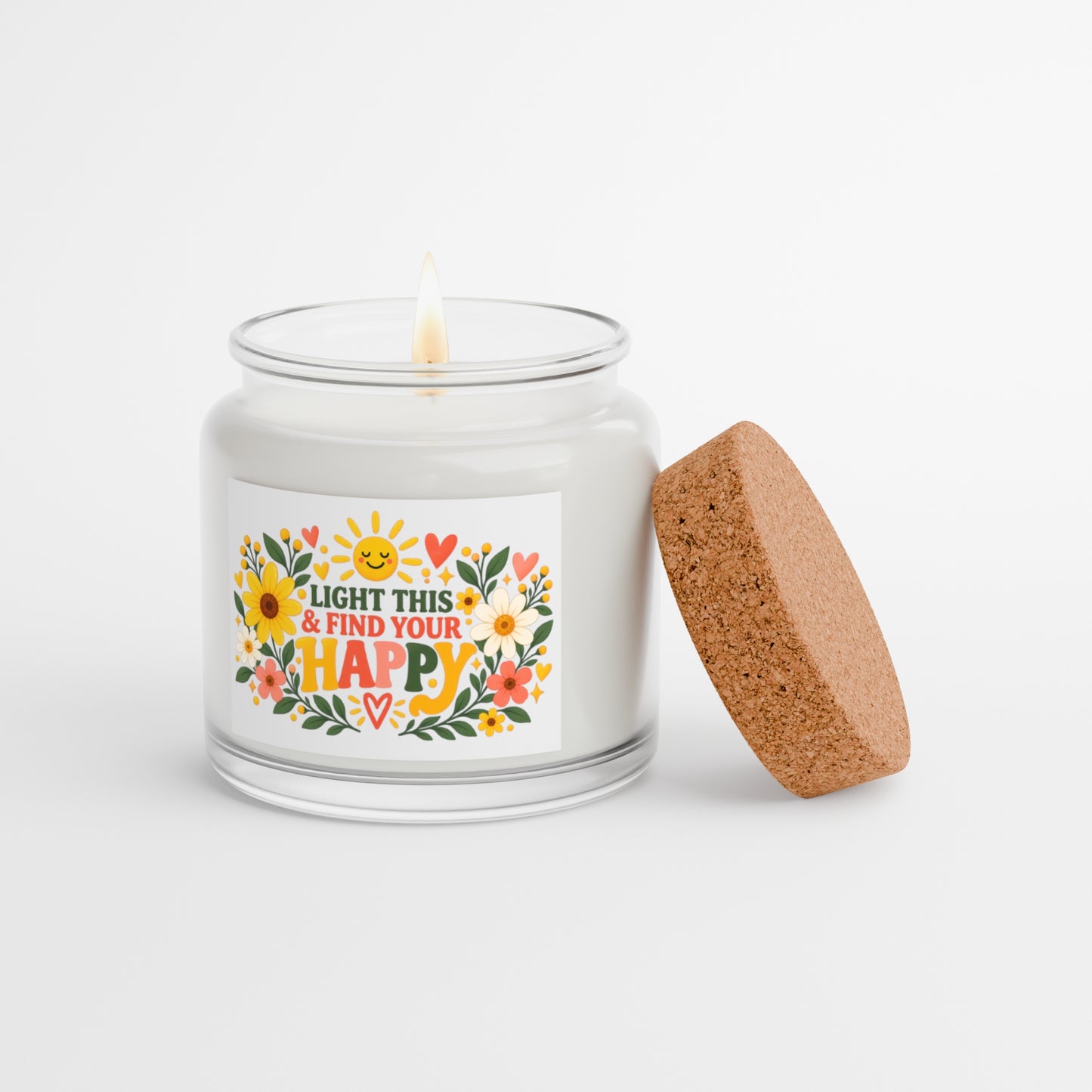 A white wax candle in a clear glass jar with a single lit wick, featuring a colorful floral label that reads 'LIGHT THIS & FIND YOUR HAPPY'. A round cork stopper or lid leans against the right side of the jar on a clean white background.