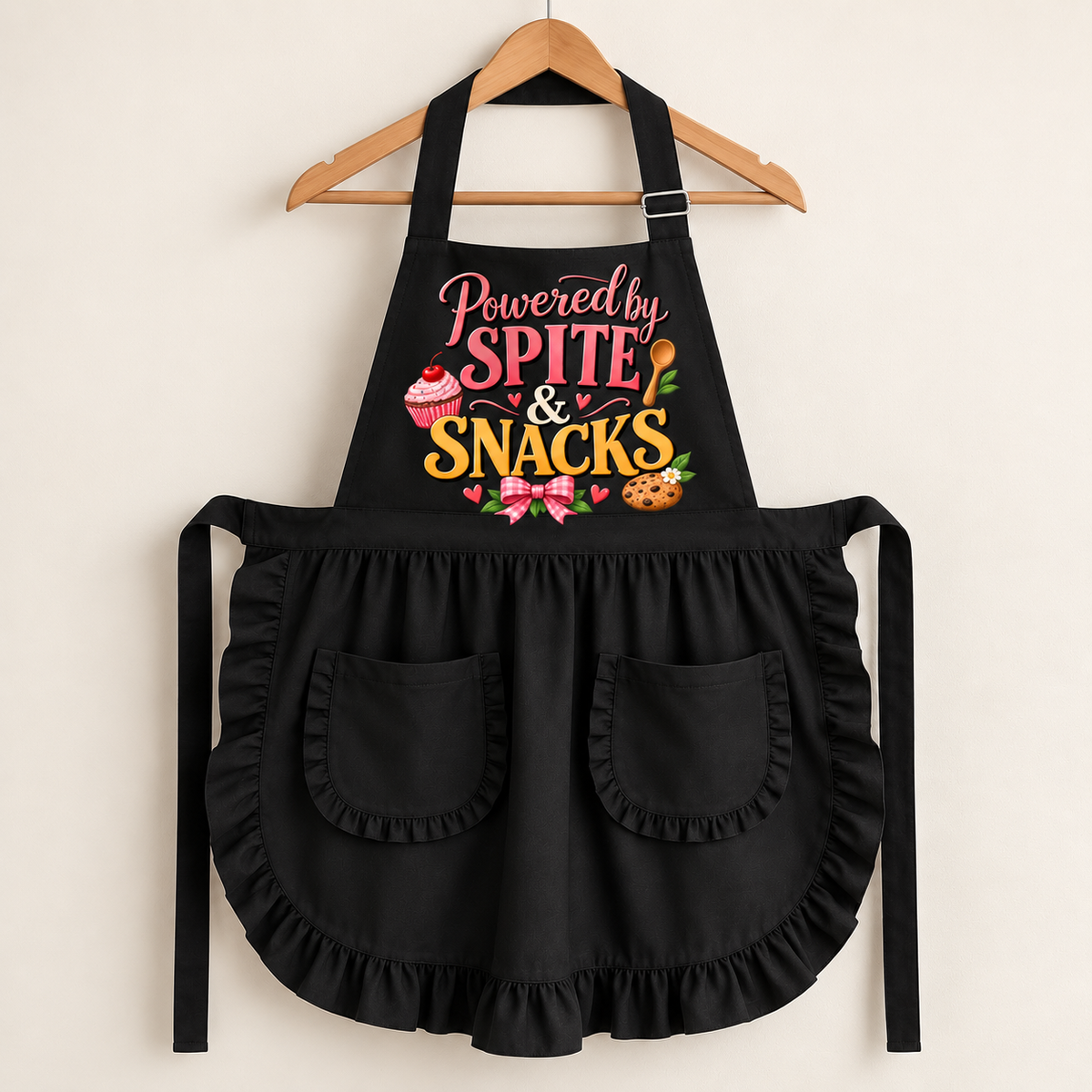 A black apron with ruffled trim and two front pockets, hanging on a wooden hanger against a light beige wall. The apron features a colorful graphic on the chest that reads 'Powered by SPITE & SNACKS' with illustrations of a cupcake, chocolate bar, cookies, and pink bows.
