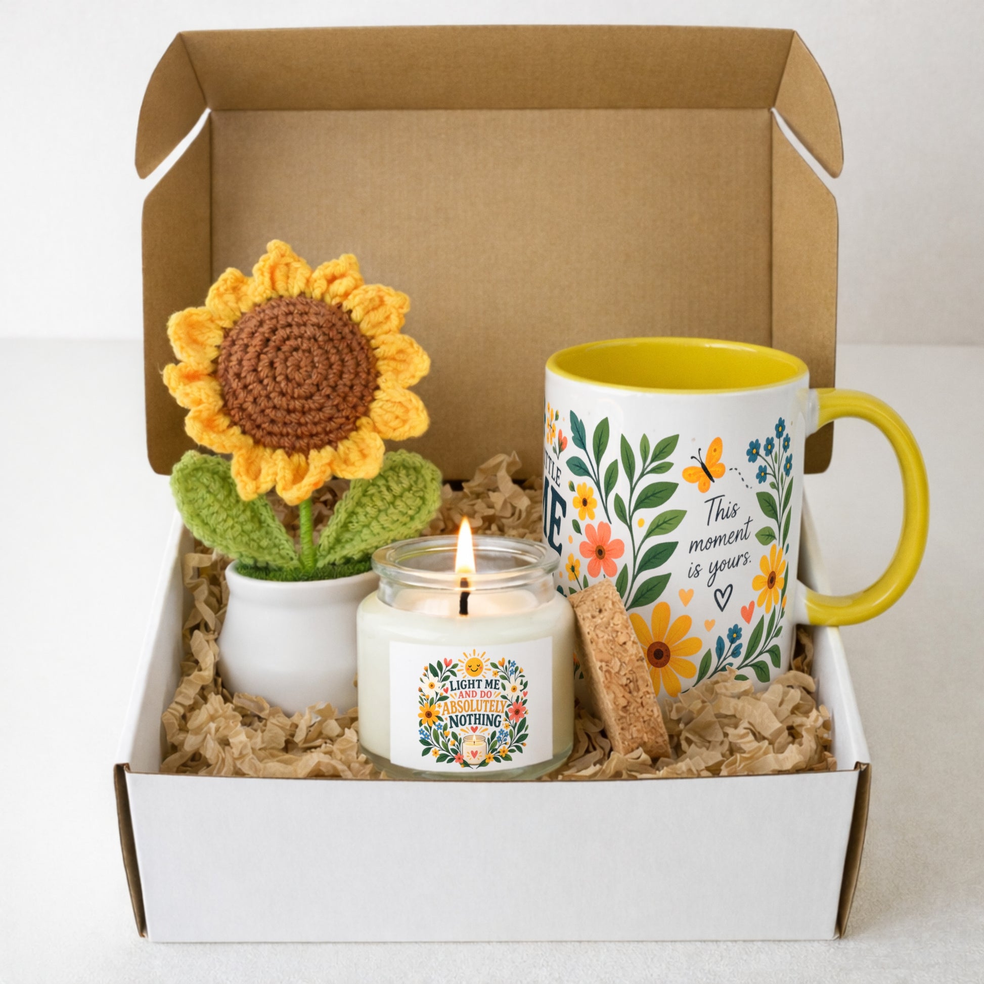 A curated gift box set containing a yellow and green crocheted sunflower in a small white pot, a white candle in a glass jar with a lit flame, a white ceramic mug with a yellow handle and interior, and a cork coaster, all arranged in brown paper shred inside an open white and brown gift box.