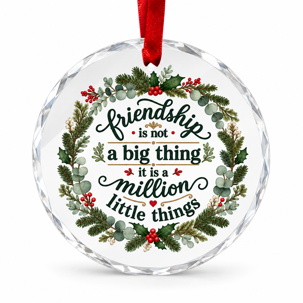 A round clear faceted glass ornament with a red hanging ribbon and printed holiday text reading 'friendship is not a big thing it is a million little things,' surrounded by a wreath of green foliage and red berries on a white background.