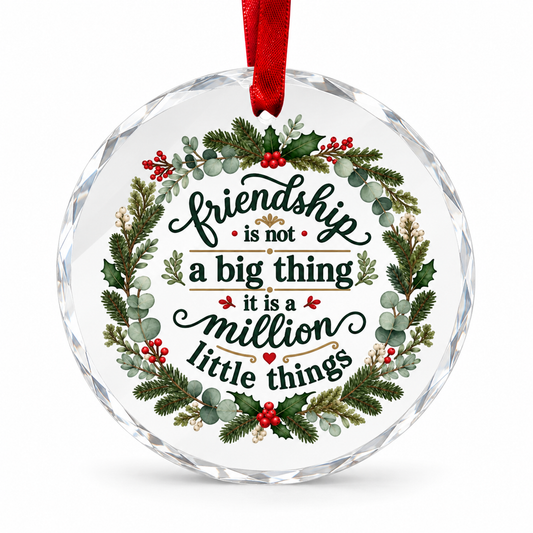 A round clear faceted glass ornament with a red hanging ribbon and printed holiday text reading 'friendship is not a big thing it is a million little things,' surrounded by a wreath of green foliage and red berries on a white background.