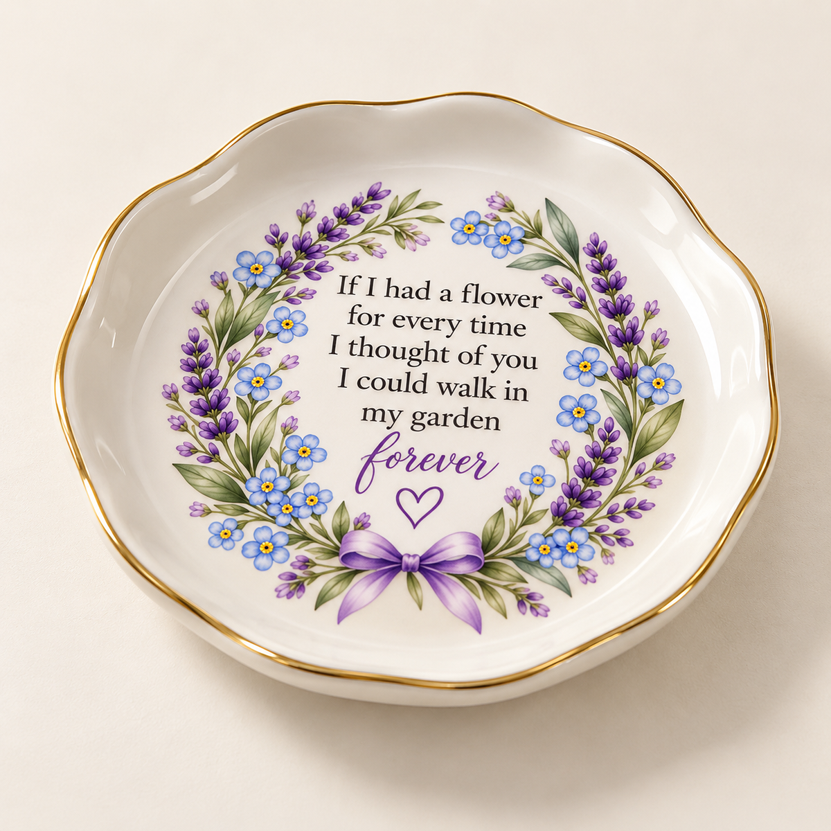 A scalloped white ceramic dish with a gold rim, featuring a floral wreath design with purple lavender, blue forget-me-nots, green leaves, and a purple bow. The center contains the printed sentimental quote, 'If I had a flower for every time I thought of you I could walk in my garden forever' with 'forever' in purple script.