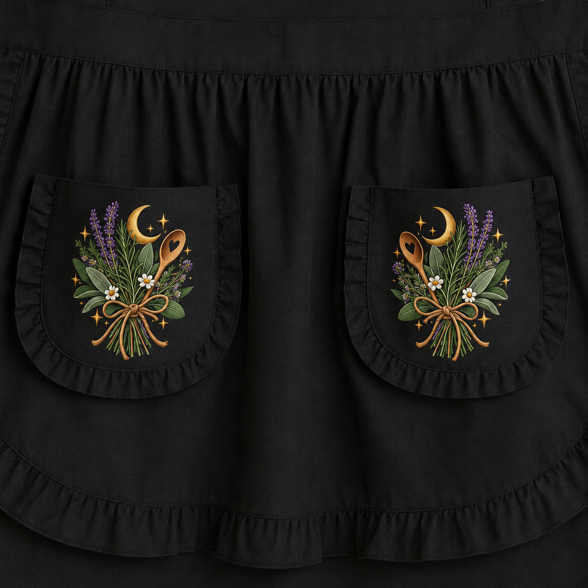 Close-up of a black apron with two ruffled front pockets, each decorated with embroidered floral and celestial motifs including purple lavender, green leaves, white flowers, a gold moon, and orange-brown utensils.