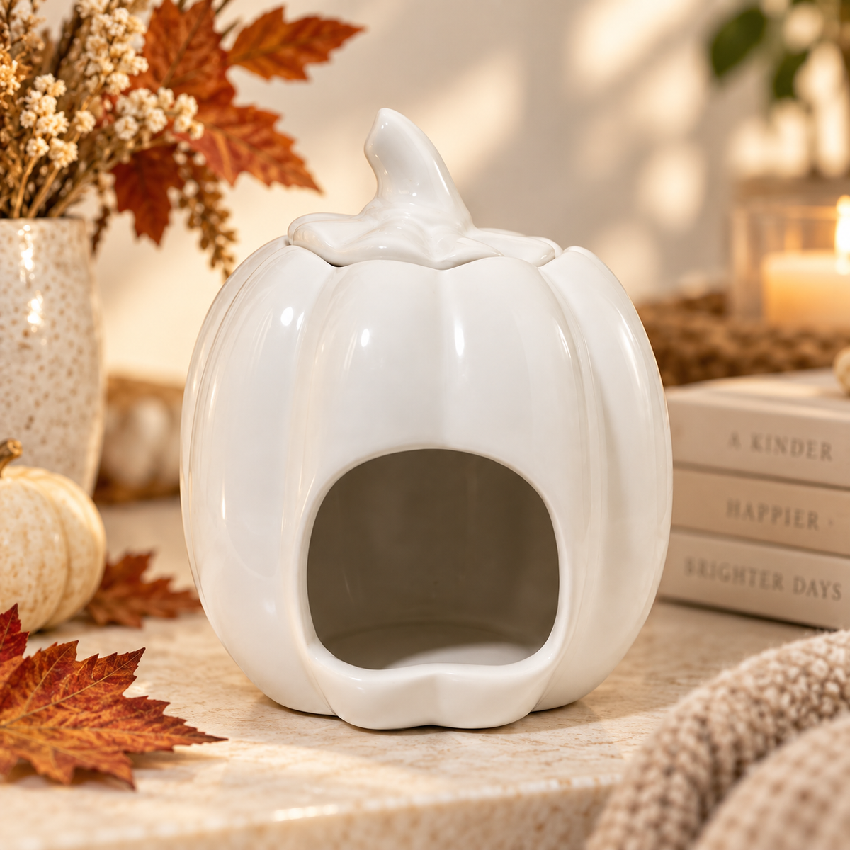 A white ceramic pumpkin-shaped candle holder with a carved opening, styled on a tabletop with autumn leaves, a small pumpkin, books, and a candle in the background.
